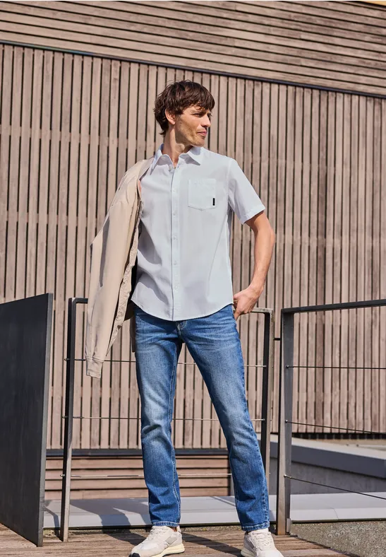 Mężczyzna w jasnoniebieskiej koszuli i dżinsach zarzuca marynarkę na ramię przed nowoczesną elewacją.