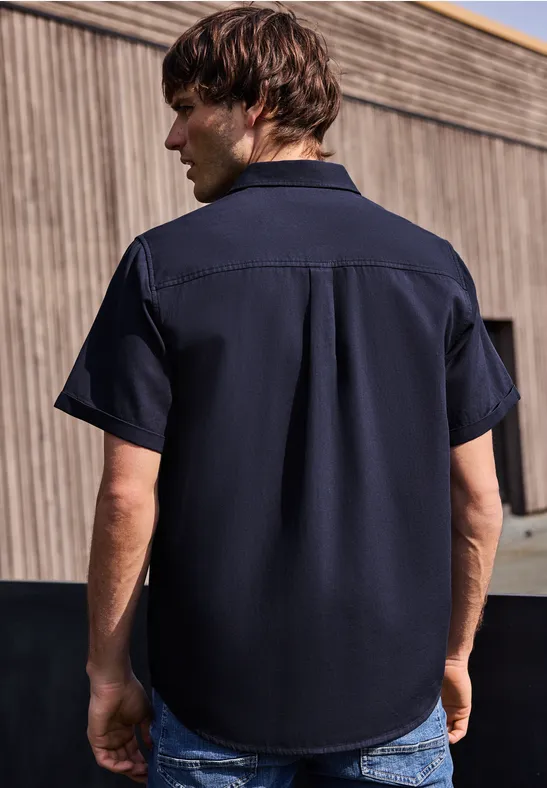 Un homme de dos porte une chemise à manches courtes bleu foncé devant une façade en bois.