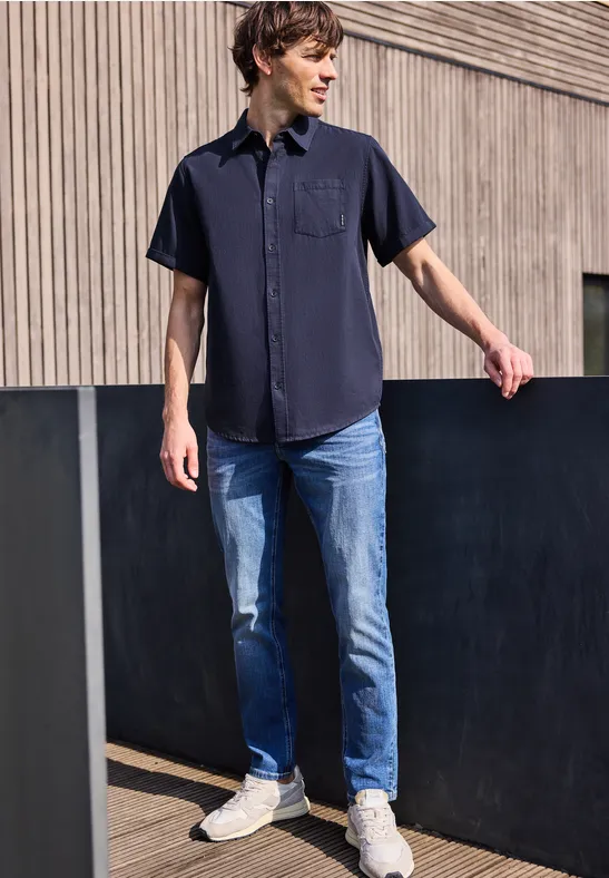 Un homme se tient devant une façade en bois, vêtu d'une chemise sombre, d'un jean bleu et de baskets blanches.
