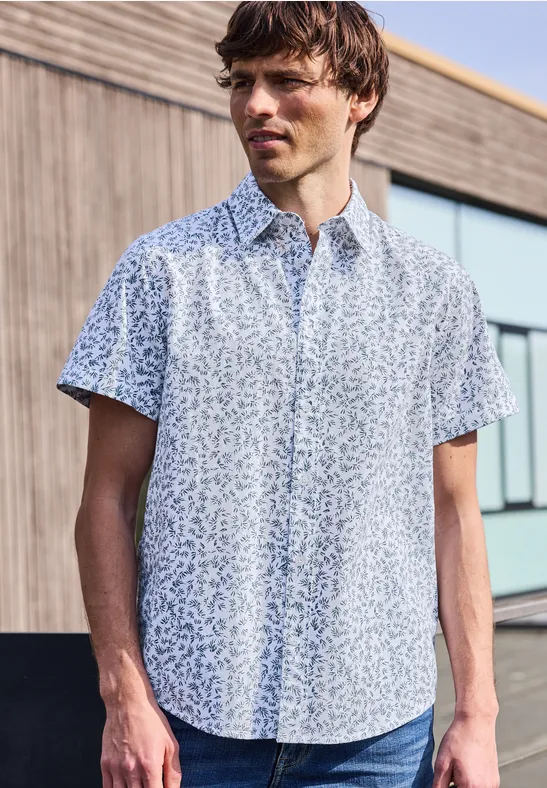 Ein Mann trägt ein kurzärmeliges, hellgemustertes Hemd und steht vor einem modernen Gebäude.