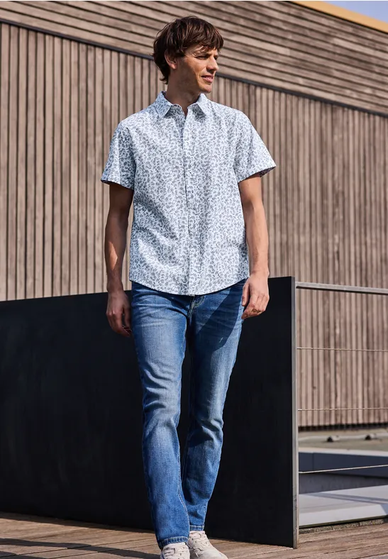 Ein Mann in gemustertem Kurzarmhemd und Jeans spaziert vor einer modernen Holzwand.