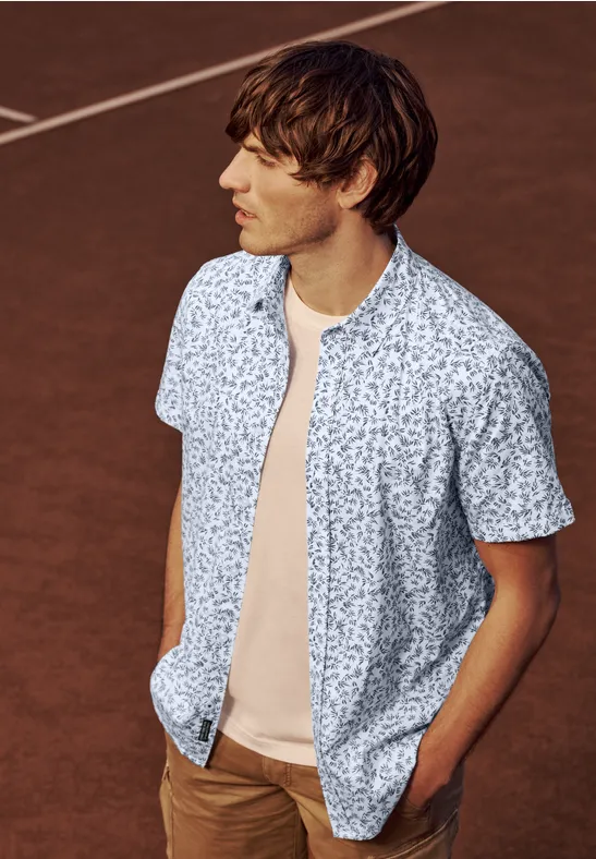 Mann in gemustertem Kurzarmhemd und T-Shirt steht entspannt auf einem Tennisplatz.