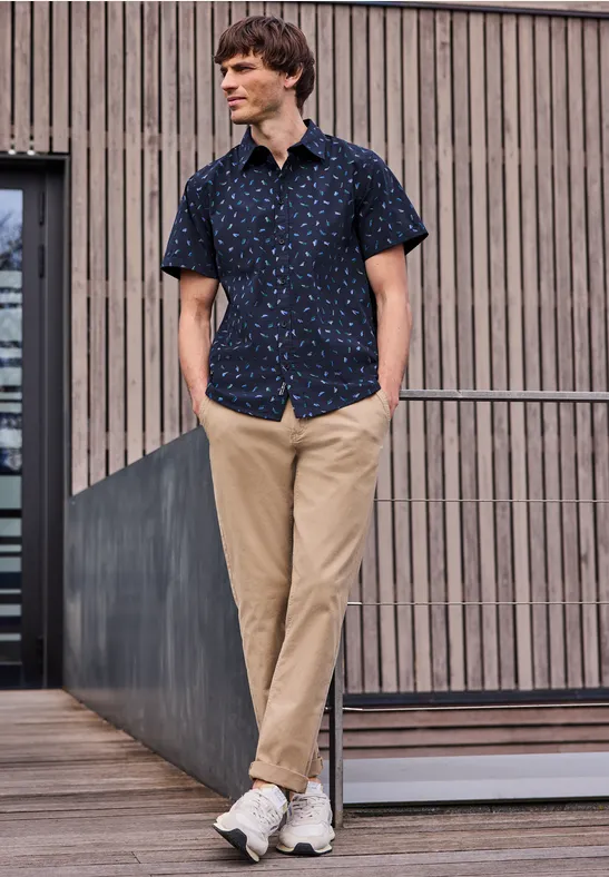Mężczyzna w wzorzystej koszuli, beżowych spodniach i sneakersach opiera się swobodnie o balustradę na tle drewnianej fasady.