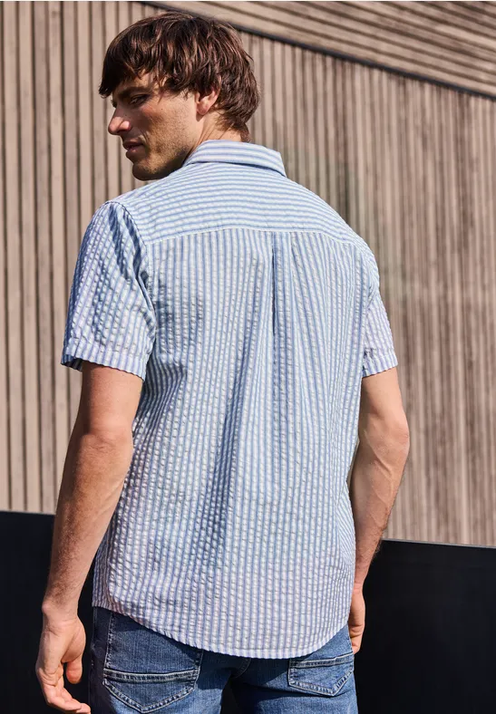 Een man staat met zijn rug naar de camera en draagt een blauw-wit geruit overhemd met korte mouwen voor een houten muur.