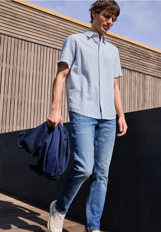 Een persoon draagt een lichtblauw kort overhemd, spijkerbroek en witte sneakers, houdt een jas vast en staat buiten.