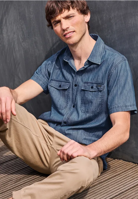 Junger Mann sitzt in legerem Jeanshemd und beigen Chinos vor einer dunklen Wand.