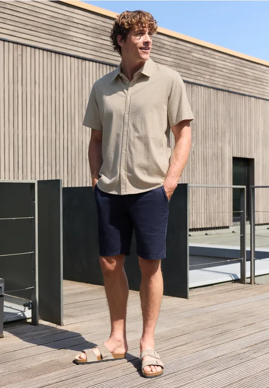 Mann in hellbeige Hemd, dunkelblauen Shorts und Sandalen steht entspannt auf Holzterrasse vor modernem Gebäude.