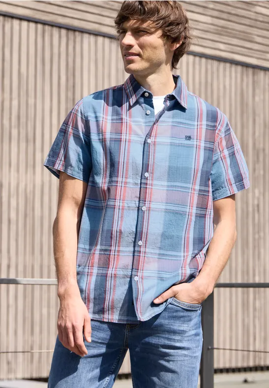 Ein Mann steht vor einer Holzwand, trägt kariertes Kurzarmhemd und Jeans, eine Hand in der Hosentasche.