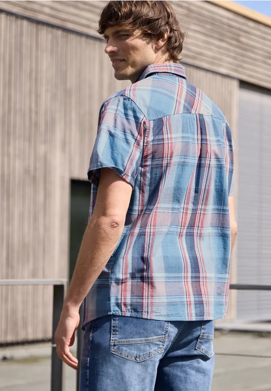 Ein Mann mit kariertem Hemd und Jeans steht vor einem Holzgebäude und schaut weg.