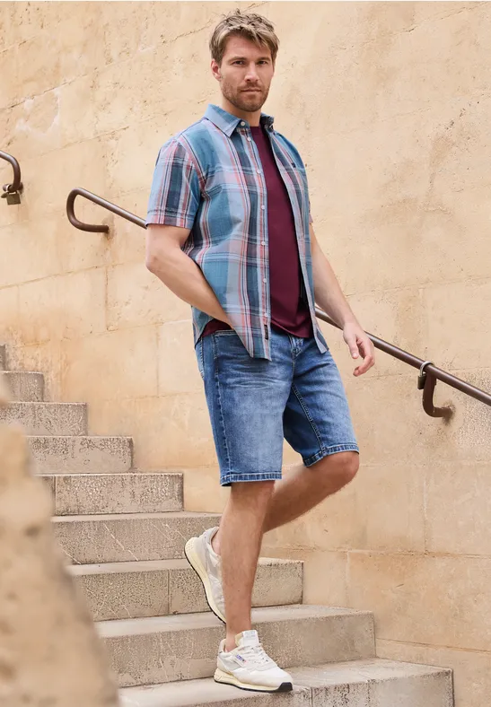 Ein Mann in Sommeroutfit steht auf einer Steintreppe und lehnt an einem Handlauf vor einer hellen Wand.