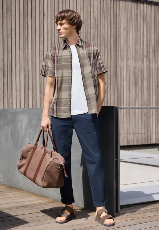 Mann in kariertem Hemd, T-Shirt, weiten Hosen und Sandalen, hält eine Reisetasche vor Holzwand.