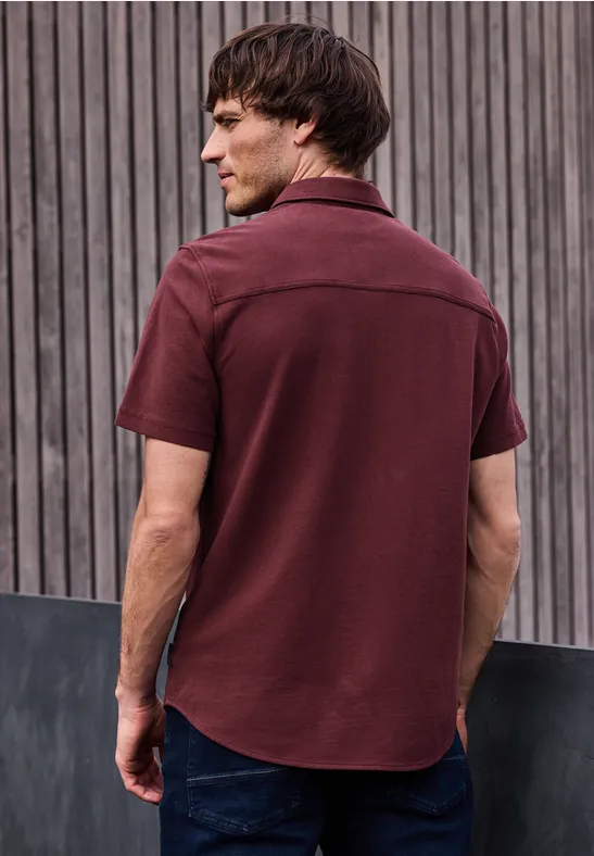 Ein Mann steht mit dem Rücken zur Kamera, trägt ein weinrotes Hemd und Jeans vor einer Holzlattenwand.