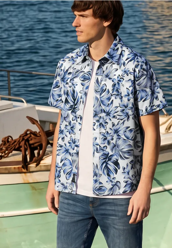 Ein junger Mann steht an einem Steg neben einem Boot und trägt Jeans, weißes T-Shirt und blau-weißes Blumenhemd.