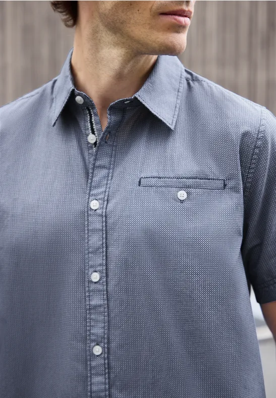 Mann mit kurzärmeligem hellblauem Jeanshemd und Brusttasche vor holzvertäfeltem Hintergrund