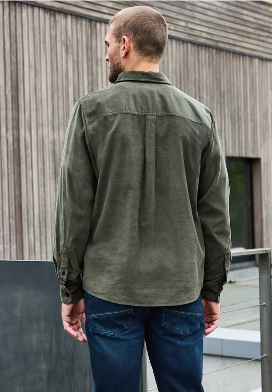 Man van achteren in groen corduroy overhemd en jeans voor een houten gevel.