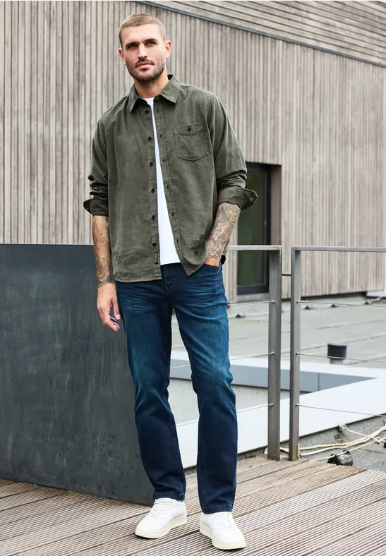 Man in kaki overshirt, wit T-shirt, blauwe jeans en witte sneakers op een dakterras.