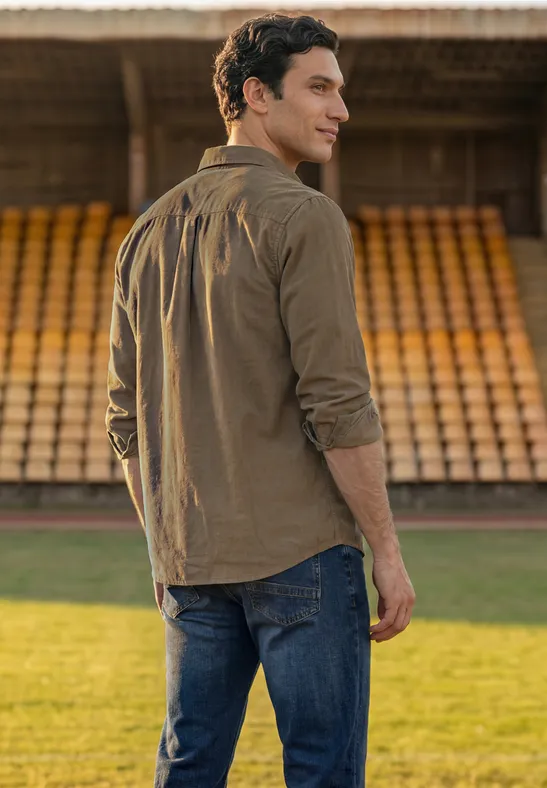 Ein Mann in braunem Hemd und Jeans steht auf einem Sportfeld und blickt über die Schulter zu den leeren Tribünen.