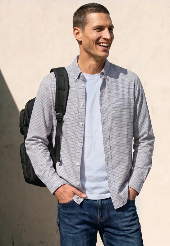 Ein lächelnder Mann in Jeans, weißem T-Shirt und hellgrauem Hemd trägt einen schwarzen Rucksack vor einer hellen Wand.