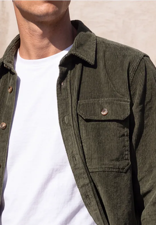 Een man draagt een olijfgroene corduroy jack over een wit T-shirt voor een lichte muur.