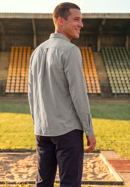 Ein Mann im grauen Hemd steht lächelnd im Stadion, Rücken zur Kamera, Blick über die Schulter.