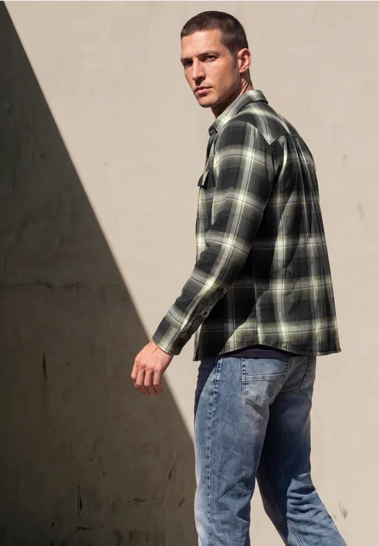 Ein Mann in kariertem grünen Hemd und Jeans geht an einer Wand entlang, Licht- und Schattenkontrast im Hintergrund.