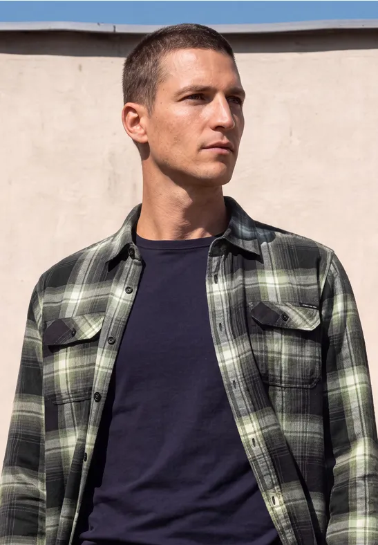 Ein Mann mit kurzem Haar trägt ein grün-schwarz kariertes Hemd und ein dunkles T-Shirt vor hellem Hintergrund.