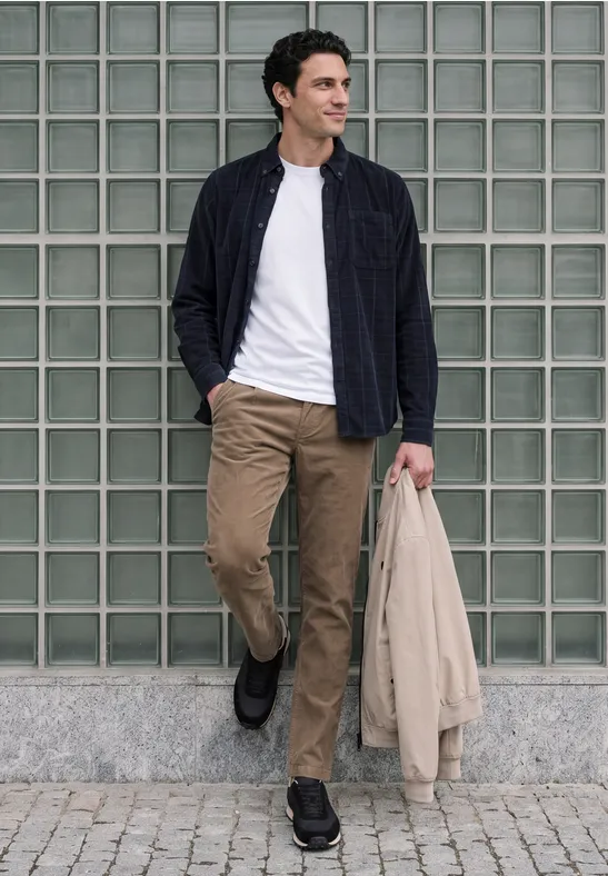 Junger Mann geht vor Glasbausteinwand, trägt hellbeige Hose, weißes Shirt, dunkle Jacke und hält hellen Mantel.