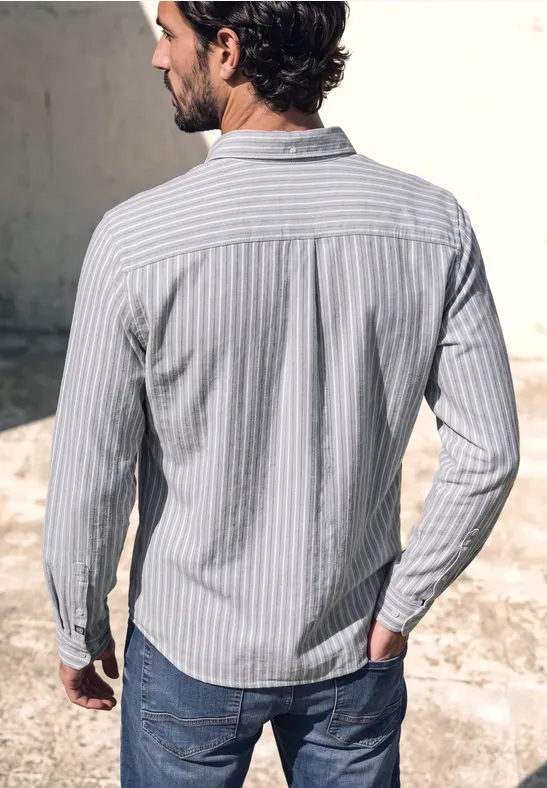 Un homme de dos, portant une chemise bleu clair rayée et un jean, se tient devant un mur gris.