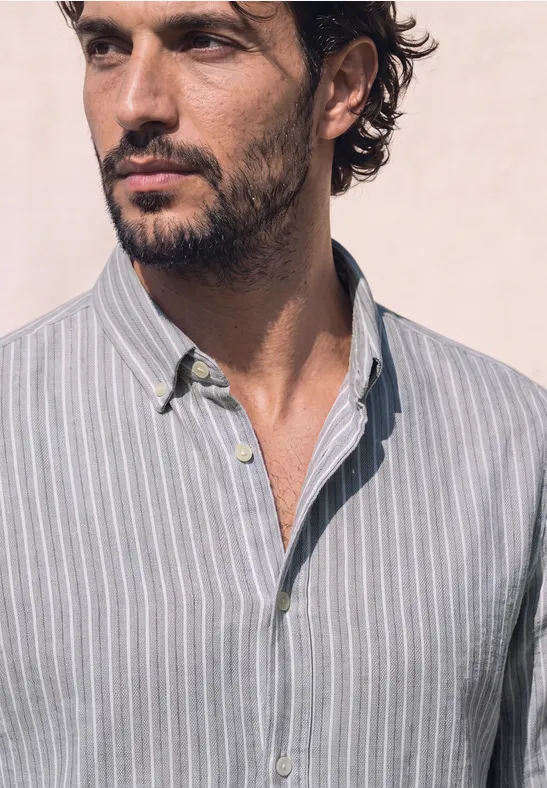 Un homme aux cheveux bruns ondulés et à la barbe discrète porte une chemise gris clair à fines rayures blanches.