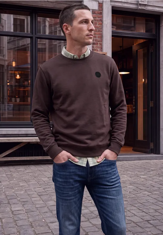Ein Mann in braunem Pullover und Jeans steht vor einem Gebäude mit großen Fenstern.