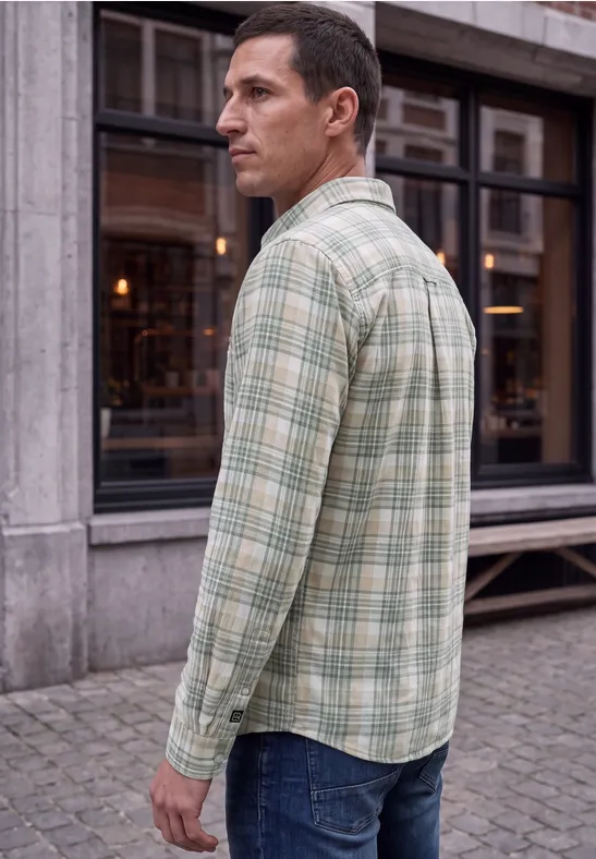 Ein Mann im Profil trägt ein hellgrünes, kariertes Hemd und Jeans, stehend auf dem Gehweg vor einem Gebäude.