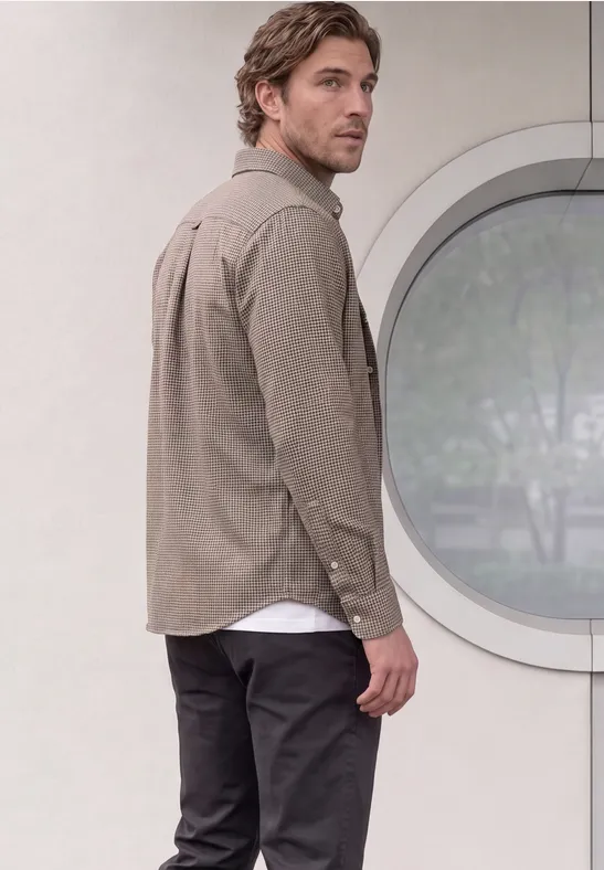 Ein Mann in beigem Hemd und dunkler Hose blickt über die Schulter vor einem runden Fenster in einer weißen Wand.