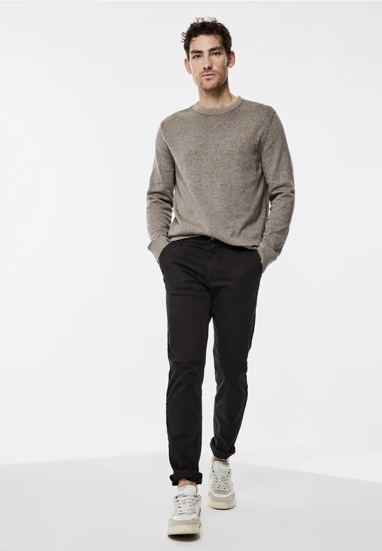 Mann in grauem Pullover, schwarzer Hose und weißen Sneakers auf weißem Hintergrund.