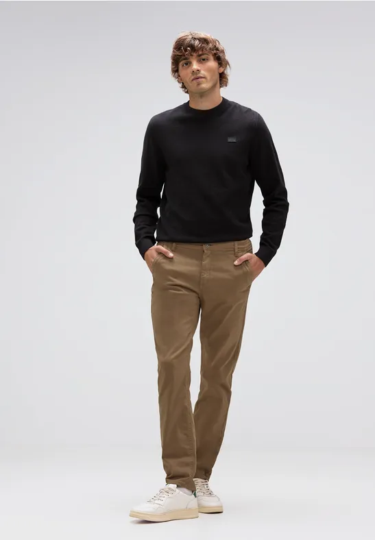 Mann trägt schwarzen Pullover, braune Hose und weiße Sneaker, steht locker.