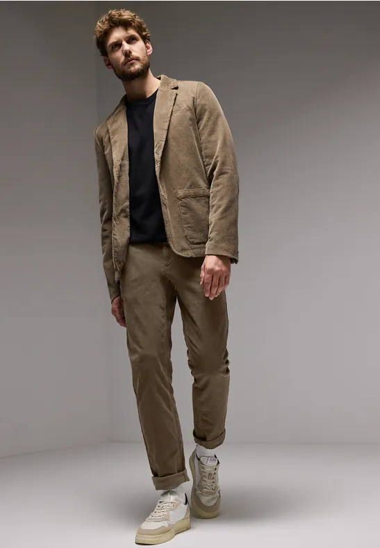 Homme en costume marron, t-shirt noir et baskets, pose sur fond gris.