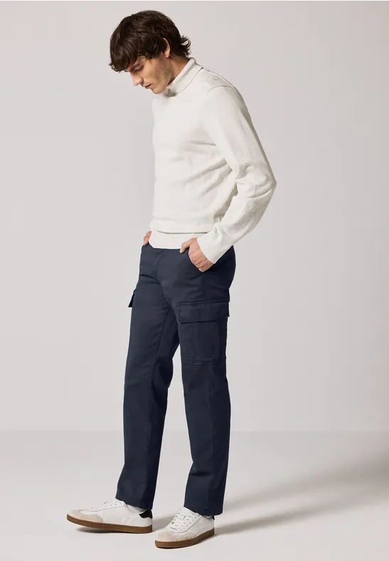 Mann mit weißem Pullover, dunkler Hose und weißen Sneakern steht seitlich.