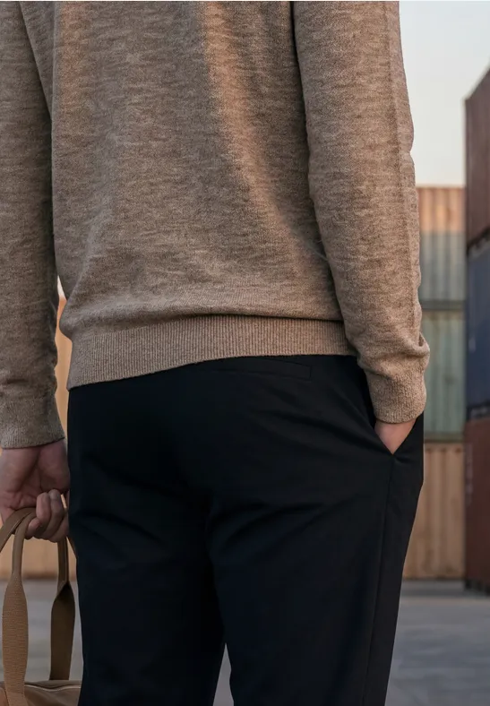 Rückansicht einer Person in grauem Pullover und schwarzer Hose, die eine Tasche hält, vor Containern.