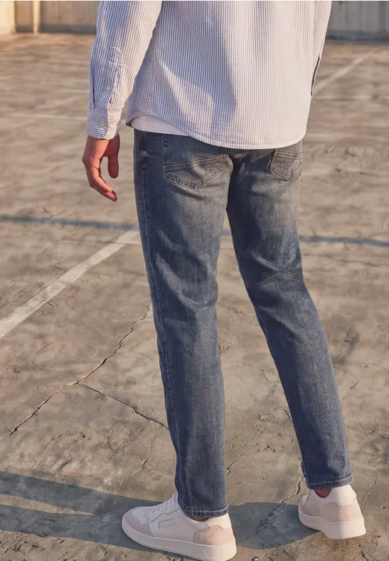 Ein Mann in Jeans und weißen Sneakern steht auf einem Außen-Sportplatz.