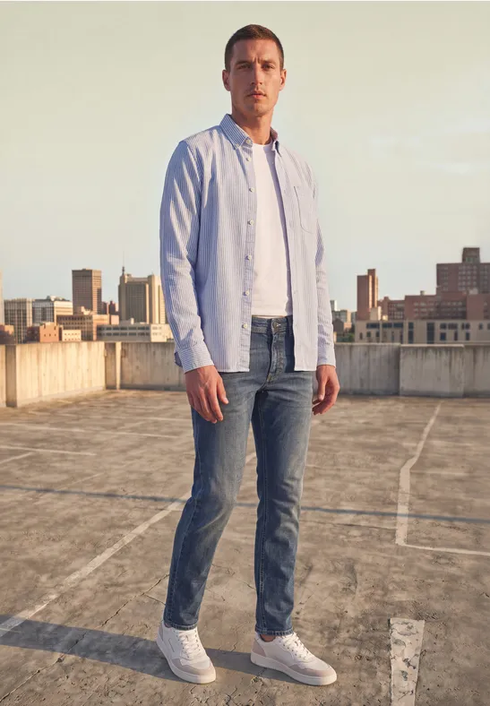 Ein Mann in Jeans, weißem T-Shirt und heller Jacke steht auf einem Parkdeck mit Stadtblick.