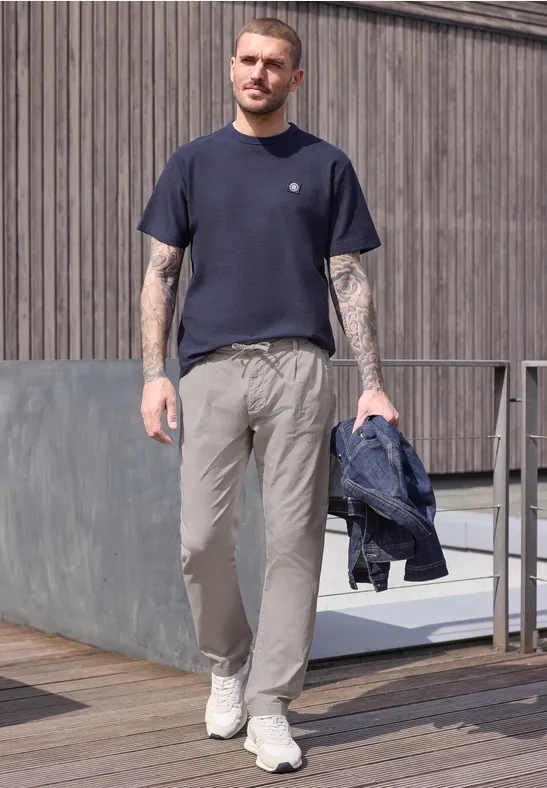 Homme tatoué sur une terrasse en bois, t-shirt bleu, pantalon gris, baskets blanches et veste à la main.