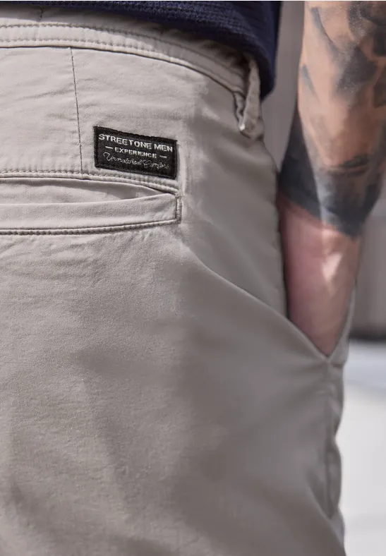 Gros plan sur un pantalon homme gris, poche arrière à écusson, main dans la poche latérale et tatouage au bras