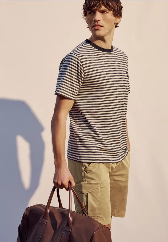 Un homme porte un t-shirt à rayures, un short kaki et tient un sac en cuir marron à la main.