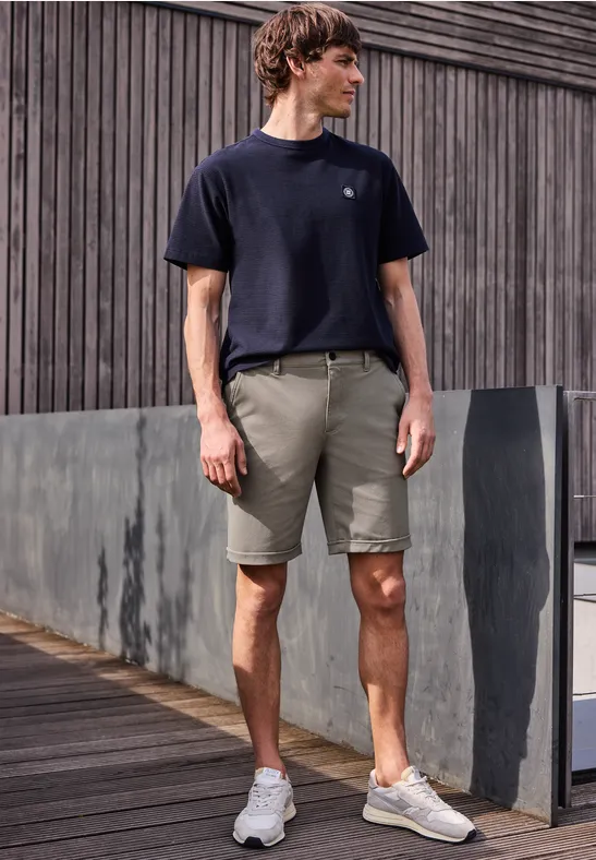 Ein Mann in schwarzem T-Shirt, grauen Shorts und weißen Sneakern steht auf einem Holzsteg vor einer grauen Wand.