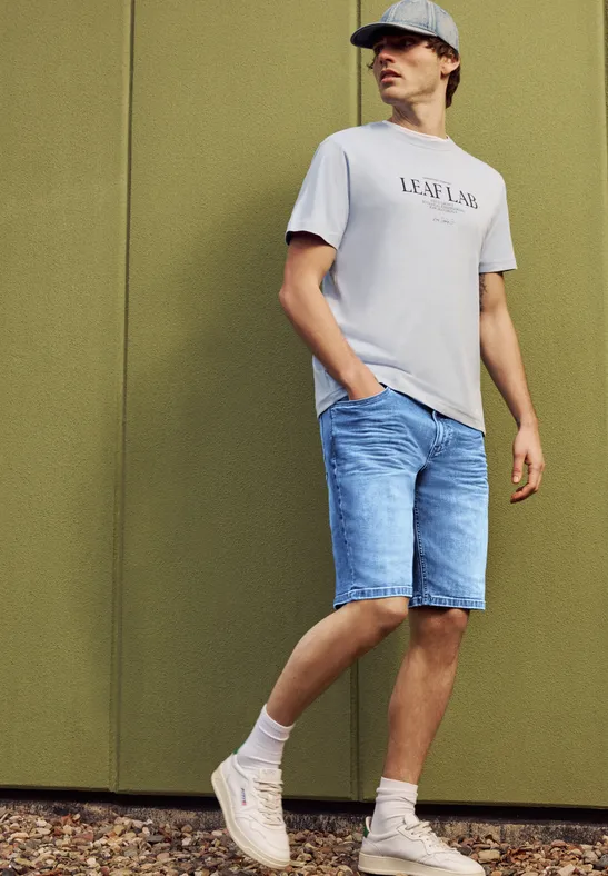 Een man in een grijs t-shirt met opschrift, blauwe jeansshort en sneakers, staand voor een groene muur.