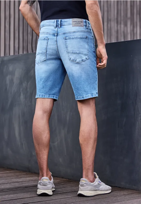 Man van achteren in lichtblauwe denimshorts, leunend tegen een hout- en betonnen muur.