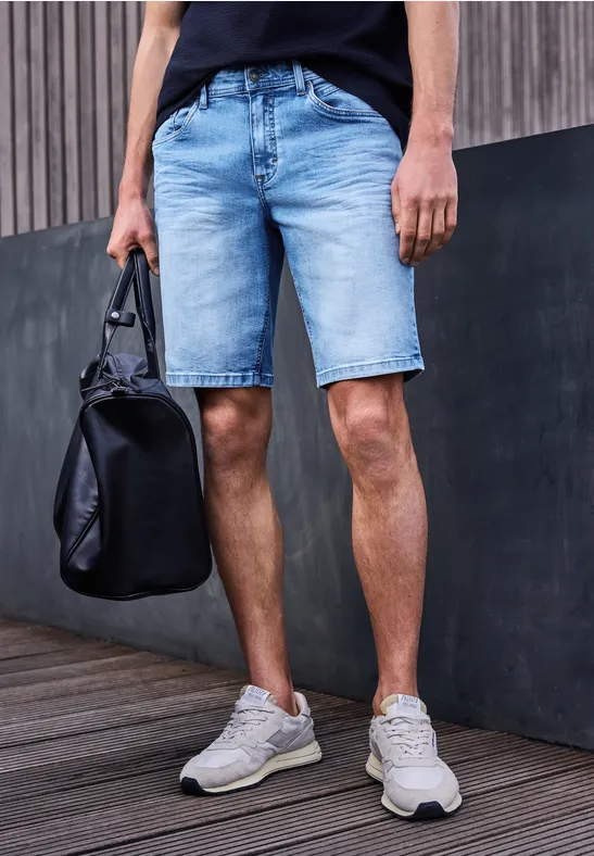 Man in jeansshorts en sneakers staat met zwarte tas aan de arm voor een muur.