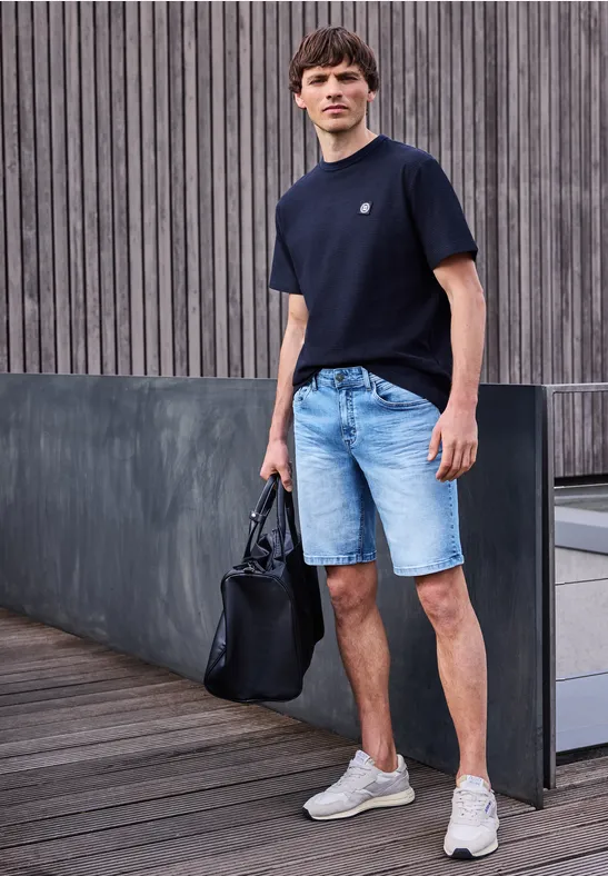 Een man in een casual outfit: zwart T-shirt, spijkershort, witte sneakers en een zwarte tas op het houten dek