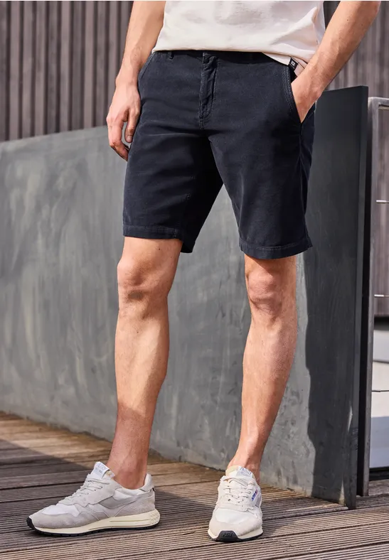 Mann in weißem T-Shirt, schwarzen Chino-Shorts und weißen Sneakern steht auf Holzdeck vor moderner Wand.