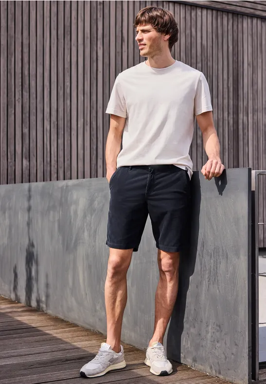 Ein Mann in weißem T-Shirt und schwarzen Shorts steht entspannt auf einem Holzsteg vor einer Fassadenwand.