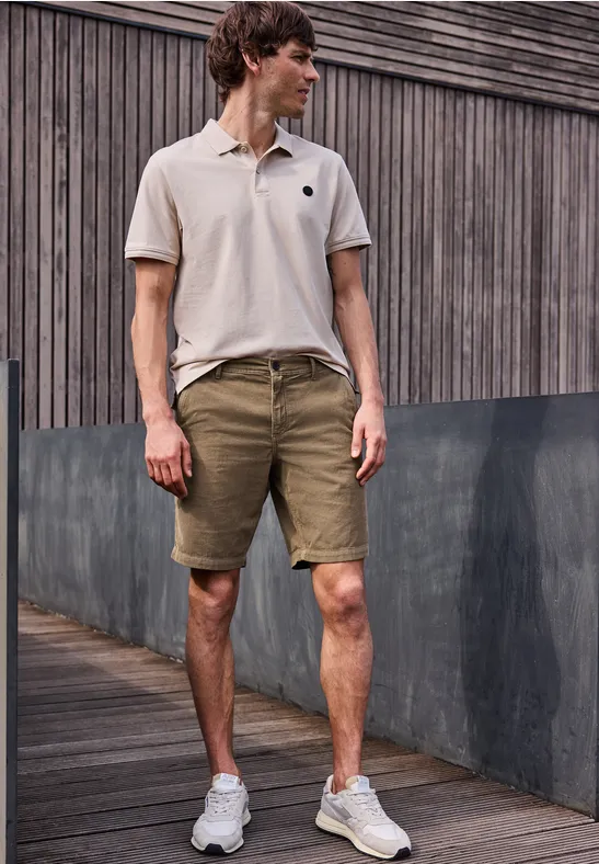 Ein Mann steht vor einer Holzfassade und trägt ein helles Polo, grüne Shorts und weiße Sneakers.